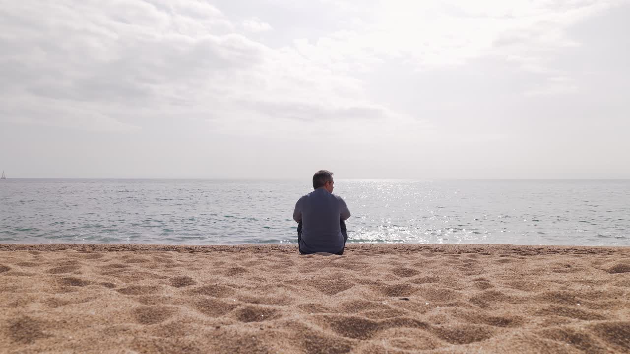 Uomo adulto seduto su una spiaggia sabbiosa, che guarda in silenzio il calmo orizzonte dell'oceano, esprimendo solitudine, riflessione e uno stile di vita sereno in riva al mare.