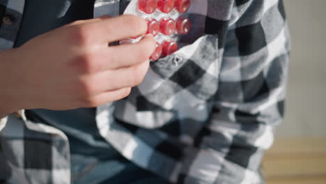 close up of man in checkered shirt reaching into pocket and pulling out red pill under bright sunlight with soft shadows on fabric and relaxed body posture suggesting self-care in urban setting