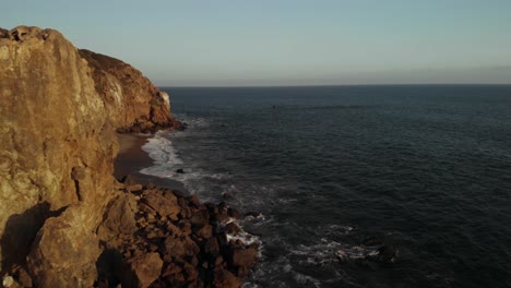 Eine-Luftaufnahme-Der-Point-Dume-Klippen-In-Malibu-In-Kalifornien,-Während-Die-Wellen-Abends-Gegen-Die-Felsen-Schlagen,-Wenn-Die-Pulsierende-Sonnenuntergang-Untergeht