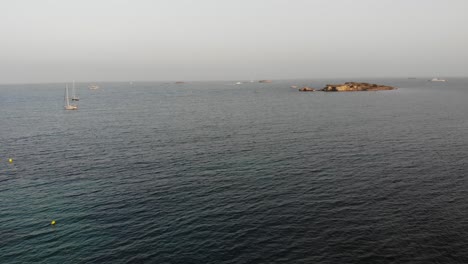 Relaxing-aerial-view-of-two-sailing-boats-and-a-small-balearic-island-in-Mediterranean-sea-with-calm-waves-on-warm-summer-afternoon-sunset-in-Eivissa,-Ibiza,-Spain