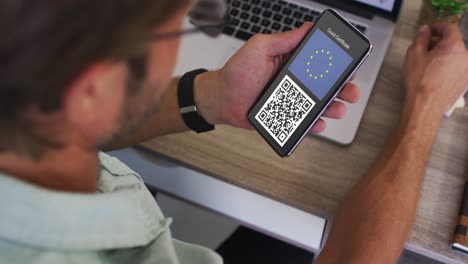 Man-at-desk-holding-smartphone-with-covid-vaccination-certificate,-eu-flag-and-qr-code-on-screen