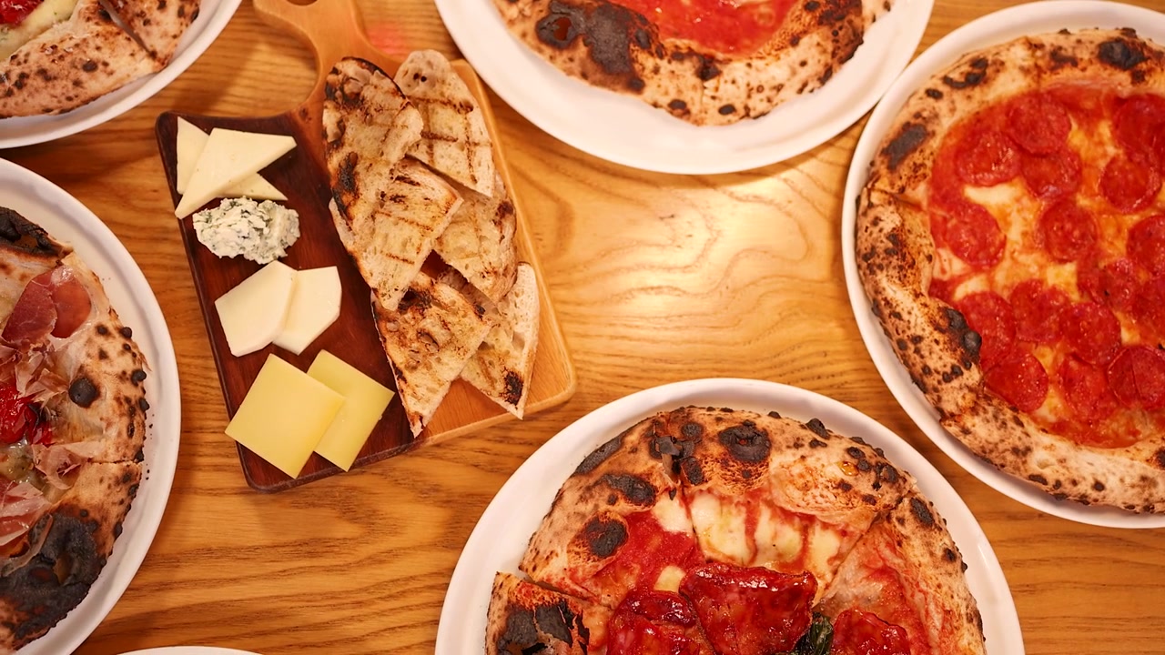 Vista superior de pizzas variadas al horno de leña y tabla de quesos en una mesa de madera