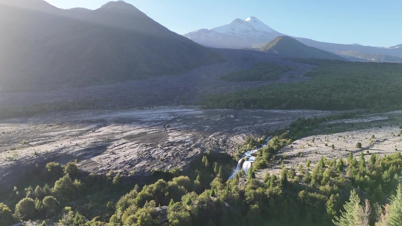 Ampia vista del Parco Nazionale Conguillío con il vulcano Llaima innevato che si erge sopra scuri campi di lava vulcanica costellati di foresta nativa. Cascate del fiume Truful Truful