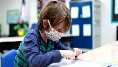 As-children-return-to-school-during-the-Corona-Virus,-Covid-19-pandemic,-a-young-boy-wearing-a-mask-attempts-to-ask-questions-and-complete-school-work