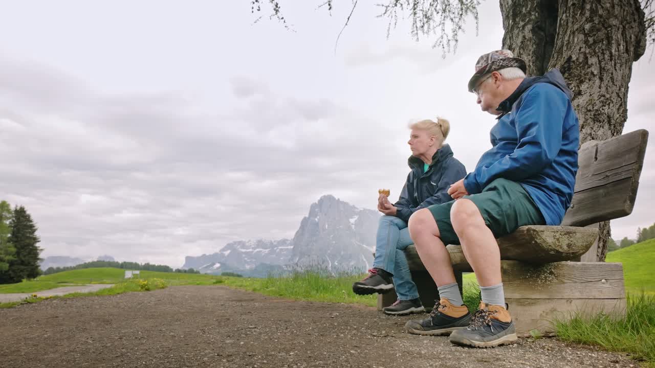 Vandrende par sidder og spiser picnic på en bænk med alpint bjerglandskab i baggrunden