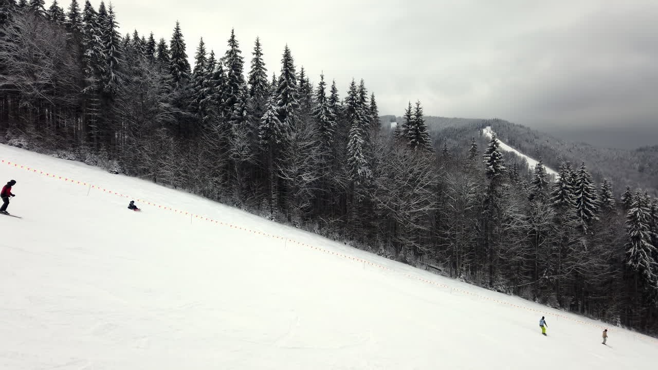 Groups of skiers navigate down snowy slopes at a mountain resort. The beautiful landscape features towering evergreen trees and a cloudy winter sky, creating a serene atmosphere