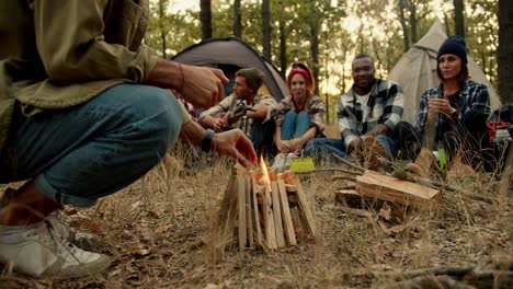 close-up shot: a man in a light green jacket makes a fire in the forest during a halt with the rest of the hikers against the backdrop of tents in a green autumn forest and withered yellow grass