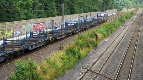 a large shipping train with empty flatbeds behind it goes down the line of railroad bordered by a graffiti-covered wall