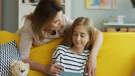 Linda-Chica-Rubia-Sentada-En-Un-Sofá-Amarillo-En-Casa-Y-Viendo-Algo-En-La-Tableta,-Luego-Su-Madre-Se-Acerca-Y-La-Abraza