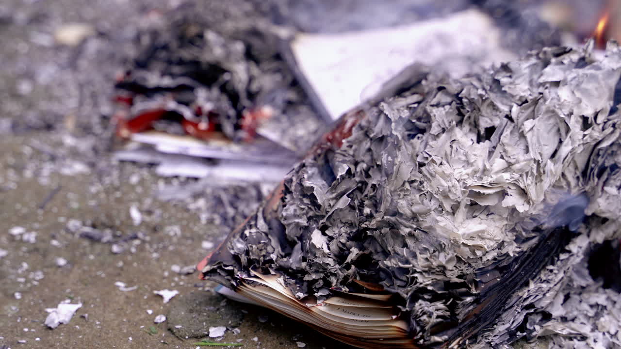 Pila di carta e libri carbonizzati che si trasformano in cenere grigia con fumo leggero. Resti fumanti di libri arancioni bruciati e cenere.