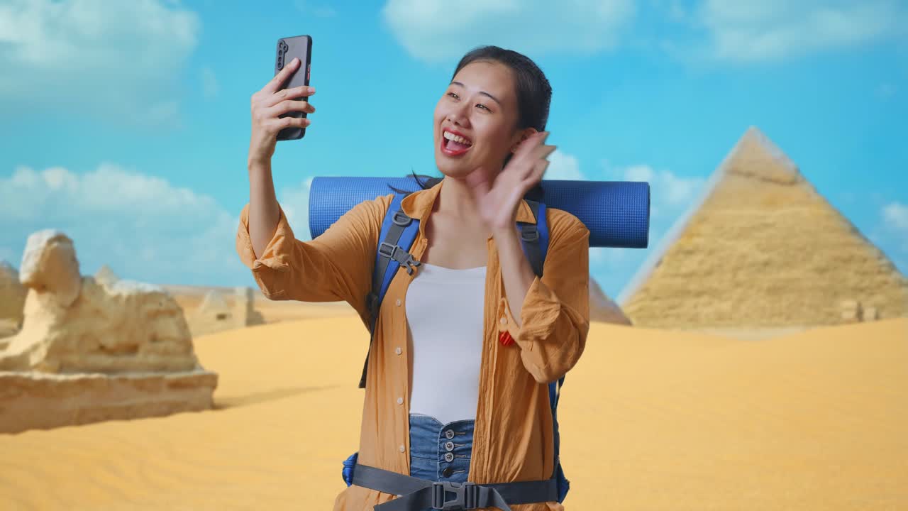 Asian Female Hiker With Mountaineering Backpack Waving Her Hand While Having A Video Call On Smartphone While Traveling In Pyramid Of Giza