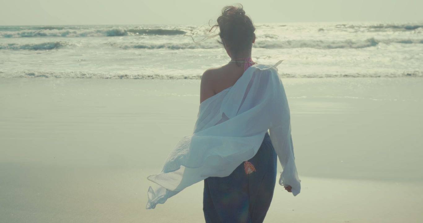 Back view of woman walking toward ocean waves on sandy beach bright sunlight calm travel lifestyle scene