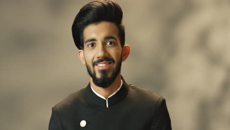 close-up view of young cheerful indian man with beard and in black jacket looking at camera and smiling cheerfully