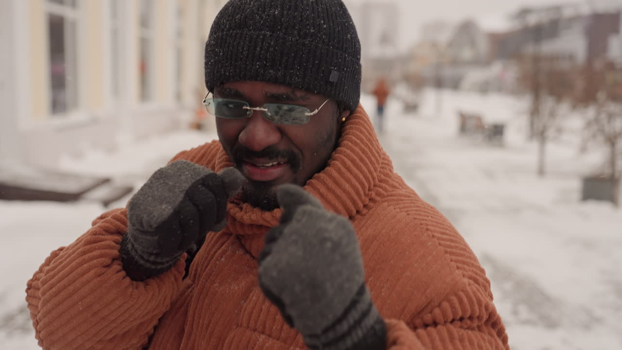 Sort mand skyggebokser på sneklædt fortov, orange sweater, strikhue, handsker, solbriller, rygsæk, dynamiske slag og hooks, legende træning, faldende sne, rå byenergi, ægte træningssession.