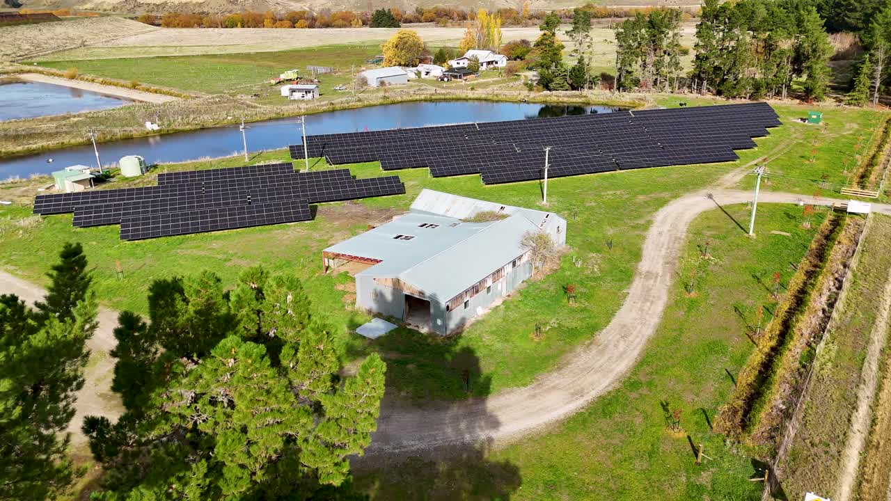 Drone footage captures a solar farm surrounded by lush greenery and a reflective pond under clear skies in Cromwell