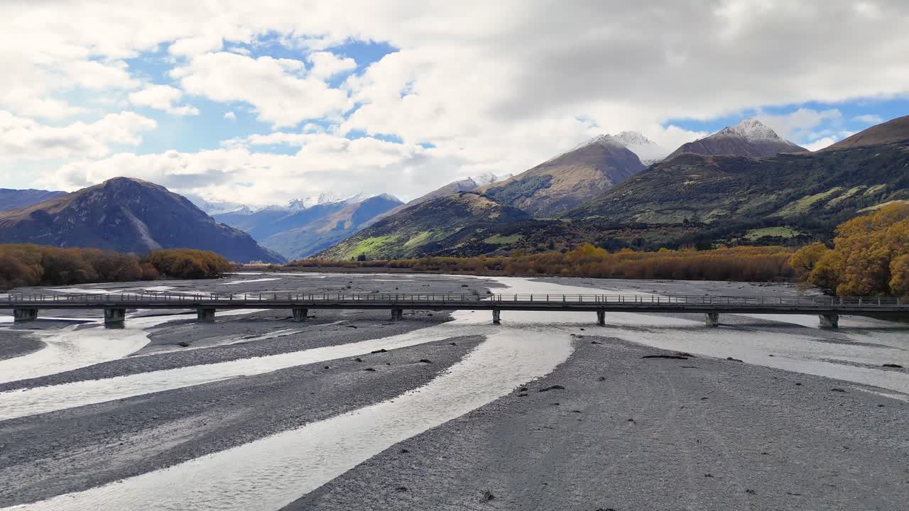 A serene view of a bridge spanning a wide, shallow riverbed with mountainous backdrop under partly cloudy skies