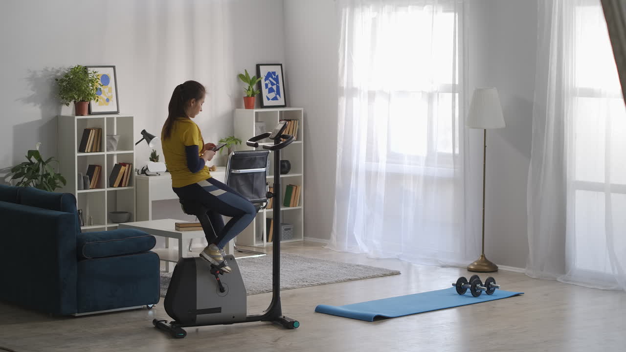 una mujer está sentada en una bicicleta estacionaria en un apartamento el fin de semana y navegando por internet con un teléfono inteligente durante el entrenamiento matutino de una joven deportiva.