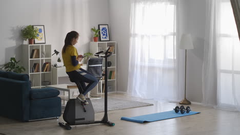 Una-Mujer-Está-Sentada-En-Una-Bicicleta-Estática-En-Un-Apartamento-Durante-El-Fin-De-Semana-Y-Navegando-Por-Internet-Con-Un-Descanso-En-El-Teléfono-Inteligente-En-El-Entrenamiento-Matutino-De-Una-Joven-Deportista