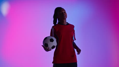 Studio-Portrait-Of-Female-Football-Or-Soccer-Player-Wearing-Team-Shirt-Holding-Ball-Under-Her-Arm-Shot-Low-Key-Against-Colourful-Mixed-Lighting-1