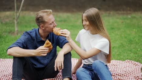 A-girl-and-a-guy-feed-each-other-a-croissant-at-a-picnic-as-a-couple.-romantic-date-happiness