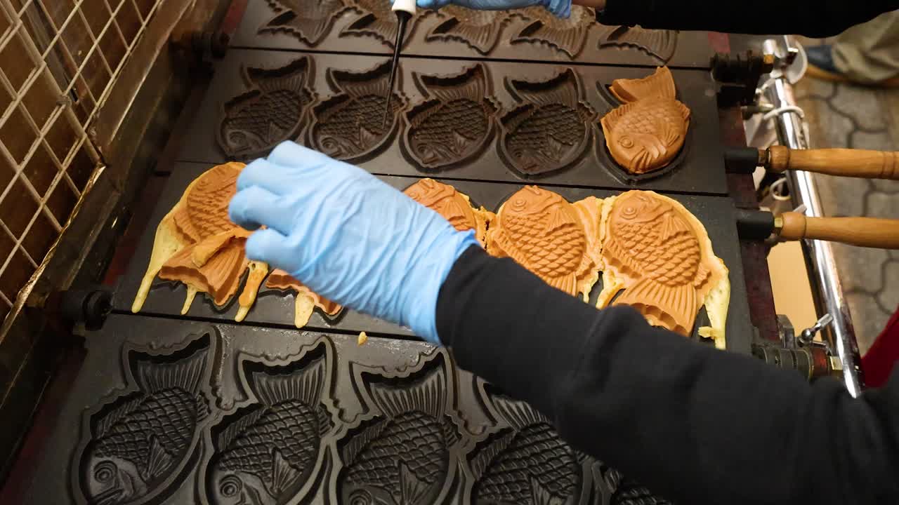 Ein Verkäufer mit blauen Handschuhen ordnet sorgfältig goldbraune Taiyaki-Kuchen auf einer heißen Grillplatte an.