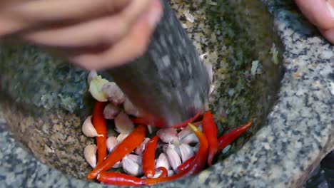 Mano-Asiática-Usando-Mortero-De-Piedra-Y-Maja-Aplastando-Ajo-Y-Ají-Rojo,-De-Cerca,-En-Cámara-Lenta