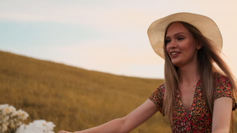 happy and cheerful girl in a hat rides a bike in a dress with flowers on the field and smiles enjoying the summer and freedom