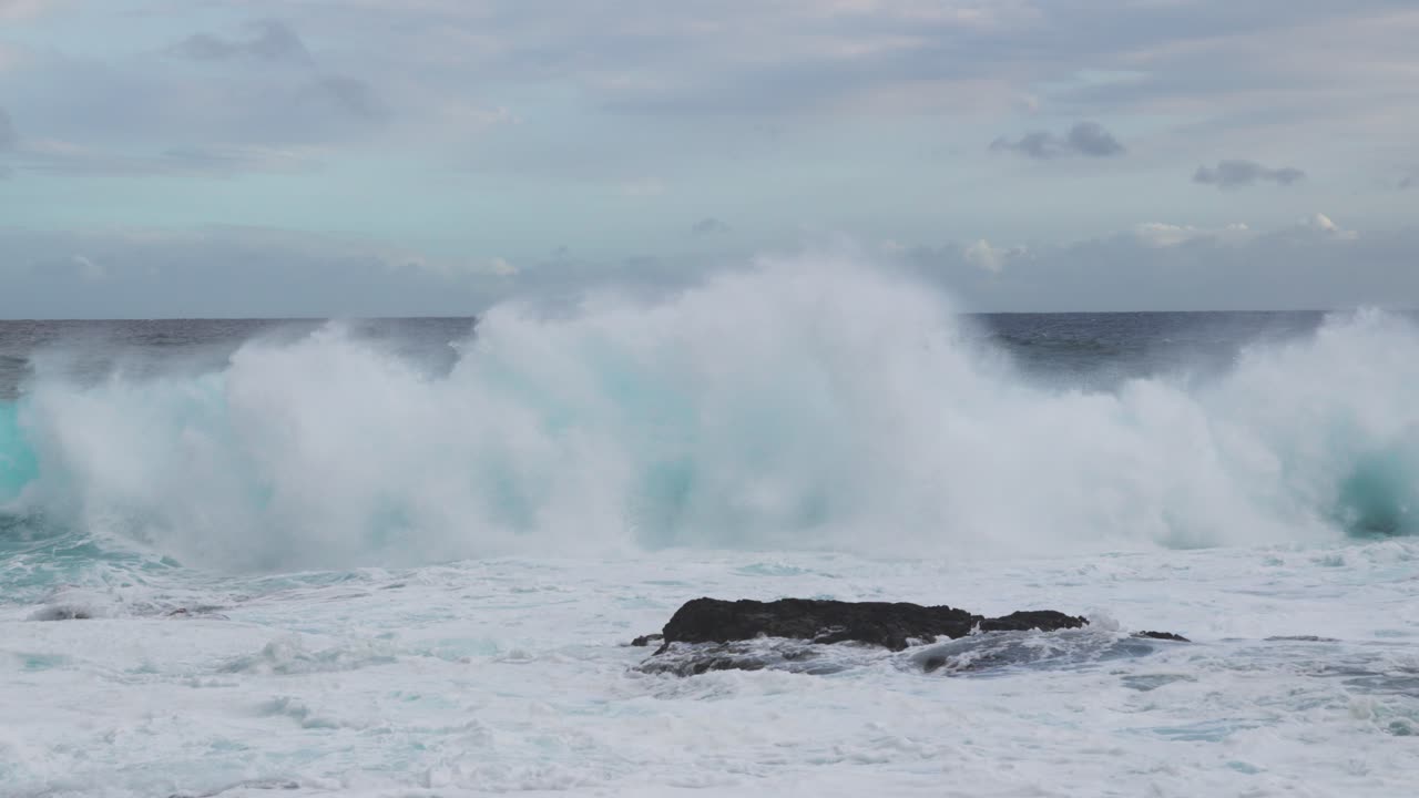 Wind-whipped teal waves rear and collapse nearshore, flinging spindrift as foamy whitewater boils around a dark rock under mottled clouds and a steady onshore breeze