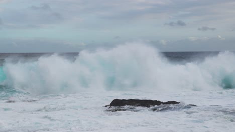 wind-whipped teal waves rear and collapse nearshore, flinging spindrift as foamy whitewater boils around a dark rock under mottled clouds and a steady onshore breeze