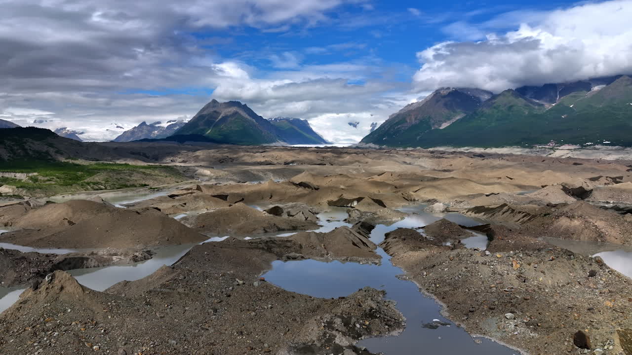 Glacial moraine landscape in Alaska. Pools of meltwater dot a gray moraine plain stretching toward distant Alaska glaciers