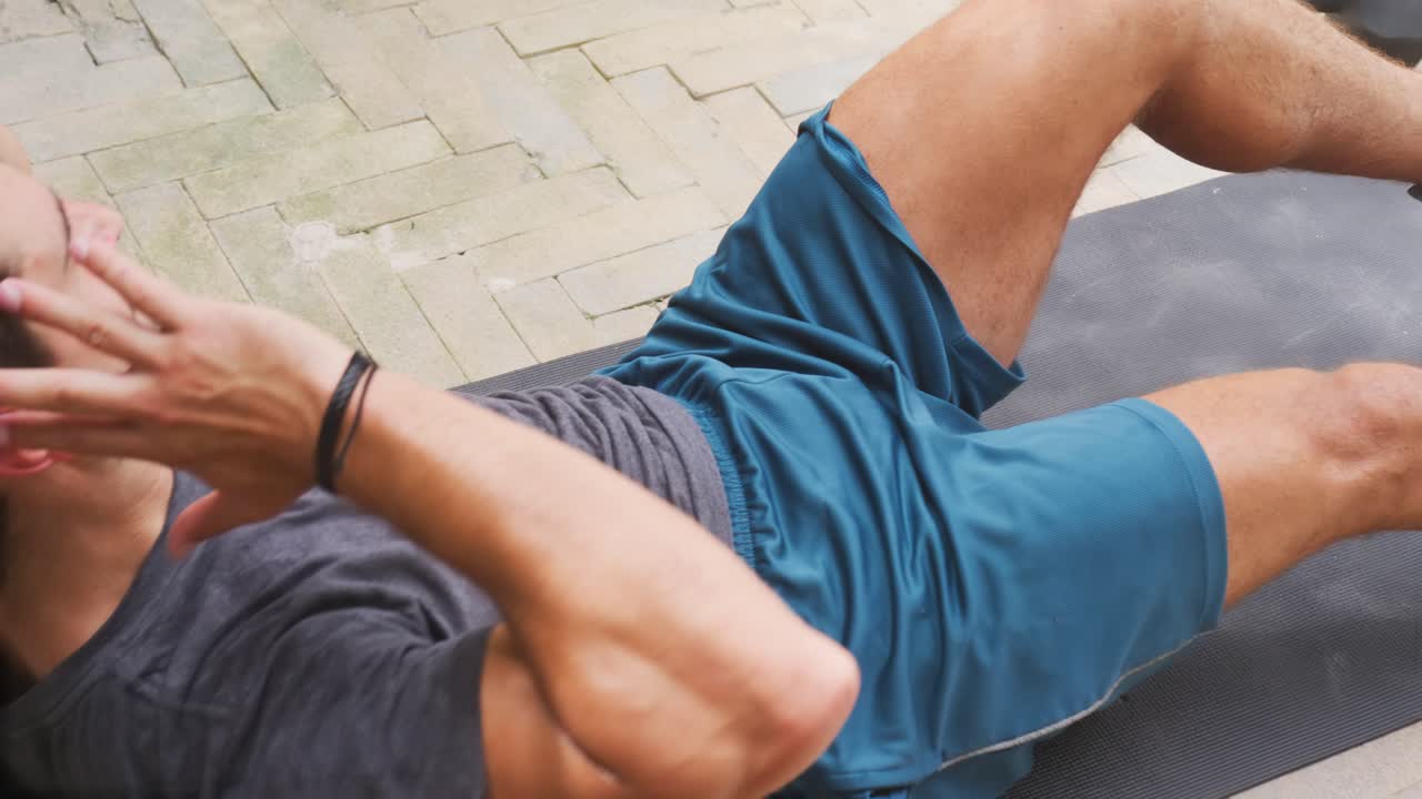Muž provádí „bike crunches“ (šlapání na kole vleže), začíná vleže na zádech na podložce na terase ve sportovním oblečení pro posílení středu těla, s činkami a lahví.