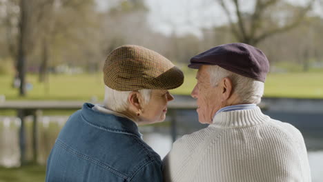 back view of an elderly couple kissing while sitting on bench in park at pond on nice autumn day