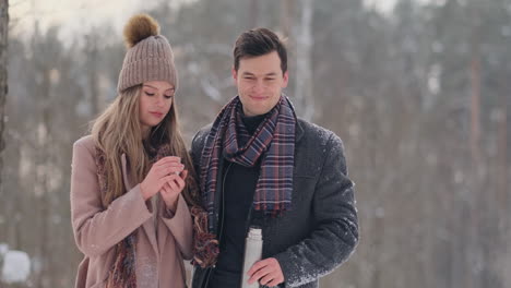 a loving couple man and woman in the winter forest drinking tea from a thermos. stylish man and woman in a coat in the park in winter for a walk. slow motion