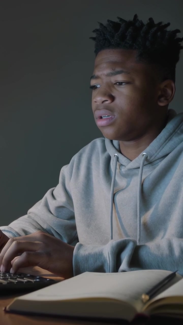 Vertical video: Typing pupil in gray hoodie reading update at desk with keys and notes rubbing head