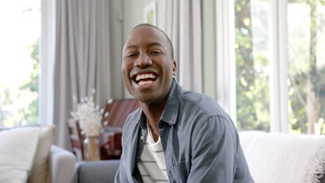 Portrait-of-happy-african-american-man-sitting-in-sunny-living-room-smiling,-slow-motion