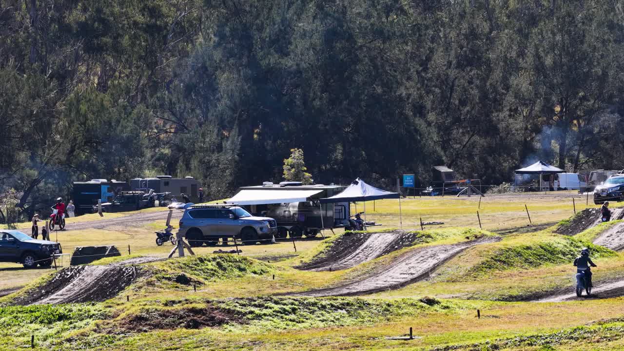 Multiple motocross riders traverse dirt track under bright daylight, surrounded by grassy fields and trees