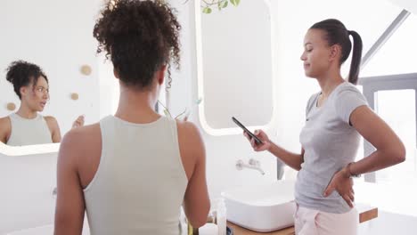 Happy-biracial-lesbian-couple-brushing-teeth-and-using-smartphone-in-sunny-bathroom,-slow-motion