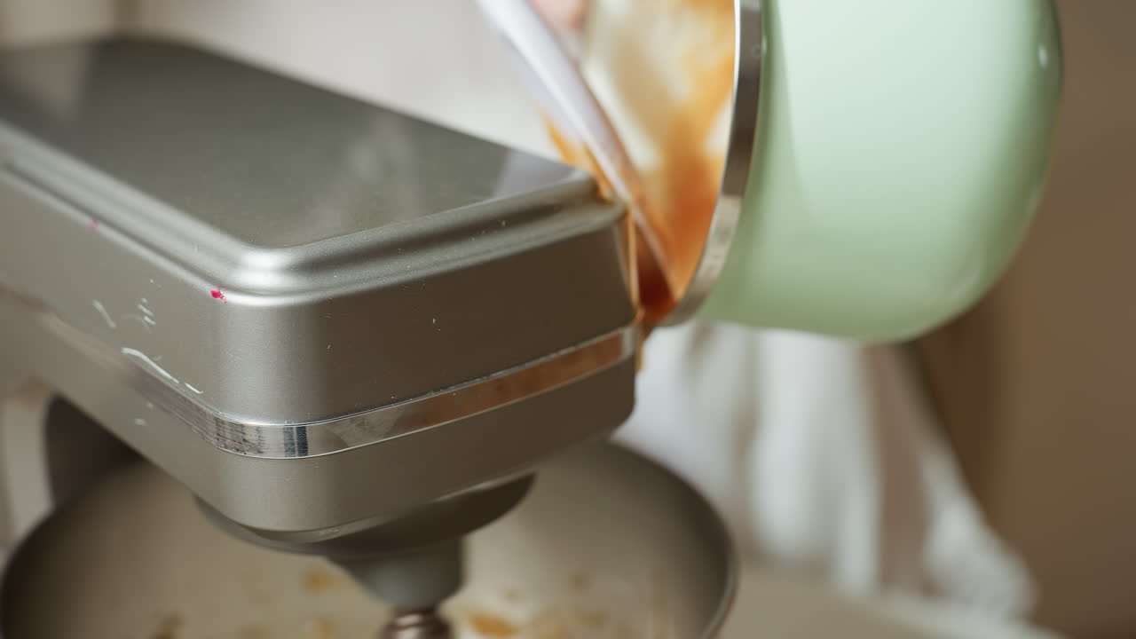 Close up of person scraping thick brown dough from green pot into electric mixer using spatula, preparing ingredients for mixing inside shiny metal bowl on countertop in bright kitchen