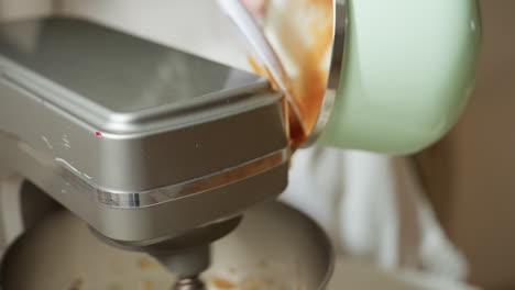 close up of person scraping thick brown dough from green pot into electric mixer using spatula, preparing ingredients for mixing inside shiny metal bowl on countertop in bright kitchen