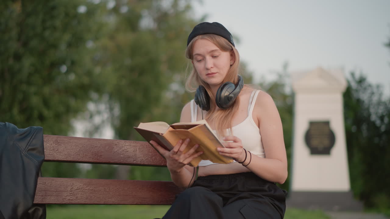 Femme blanche étudiant un livre sur un banc dans un parc verdoyant, écouteurs posés sur le cou, expression absorbée tandis qu’elle tourne les pages, veste et sac à dos à côté d’elle, douce lumière d’après-midi et lampadaire en arrière-plan