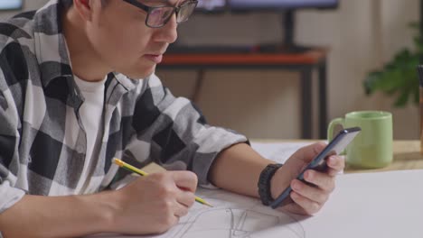close up de un hombre asiático mirando un teléfono inteligente y dibujando en papel mientras trabaja en un boceto de diseño de automóvil en la mesa en el estudio