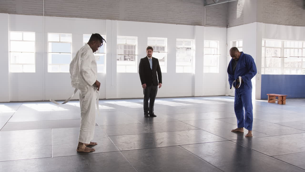 In dojo, judo practitioners tying belts with instructor observing, preparing for class