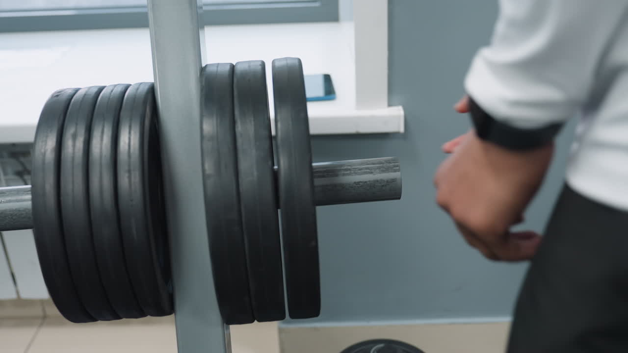 Rear view of student returning weight plate onto barbell rack in gym, showing hands guiding black iron plate back onto rack on rubber flooring during focused training session and workout preparation