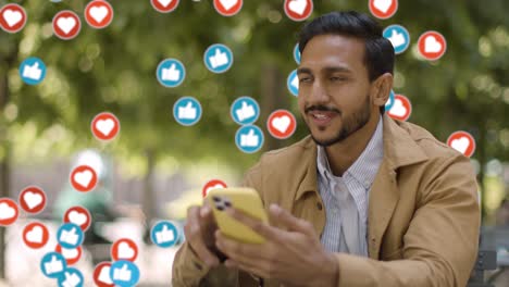 hombre sonriente sentado en una mesa al aire libre mirando su teléfono móvil con emojis de gráficos en movimiento que muestran múltiples notificaciones de redes sociales gustando y reaccionando al contenido en línea 1