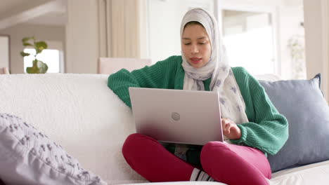 Biracial-woman-in-hijab-using-laptop-on-sofa-at-home-with-copy-space,-slow-motion