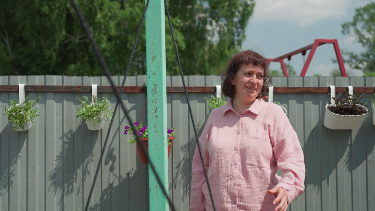 Woman Enjoying Backyard Play, Sunlit Garden Features Woman Swinging Joyfully Beside Hanging Planters, Cheerful Woman Enjoys Swinging In Sunny Garden Backyard With Lush Hanging Plants