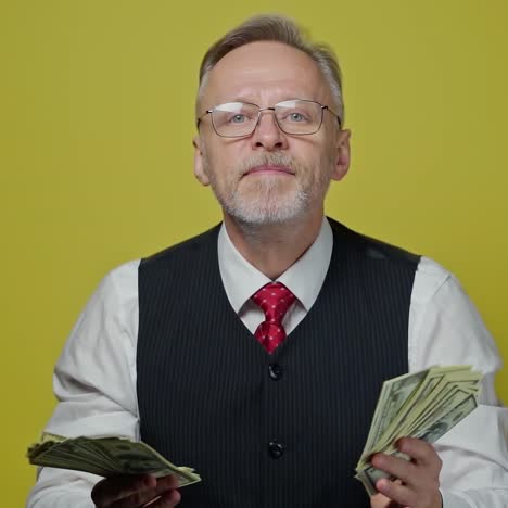 portrait of grey-haired man with dollars. smiling mature businessman in glasses waving dollar bills and touches his face with money happily. positive emotions.
