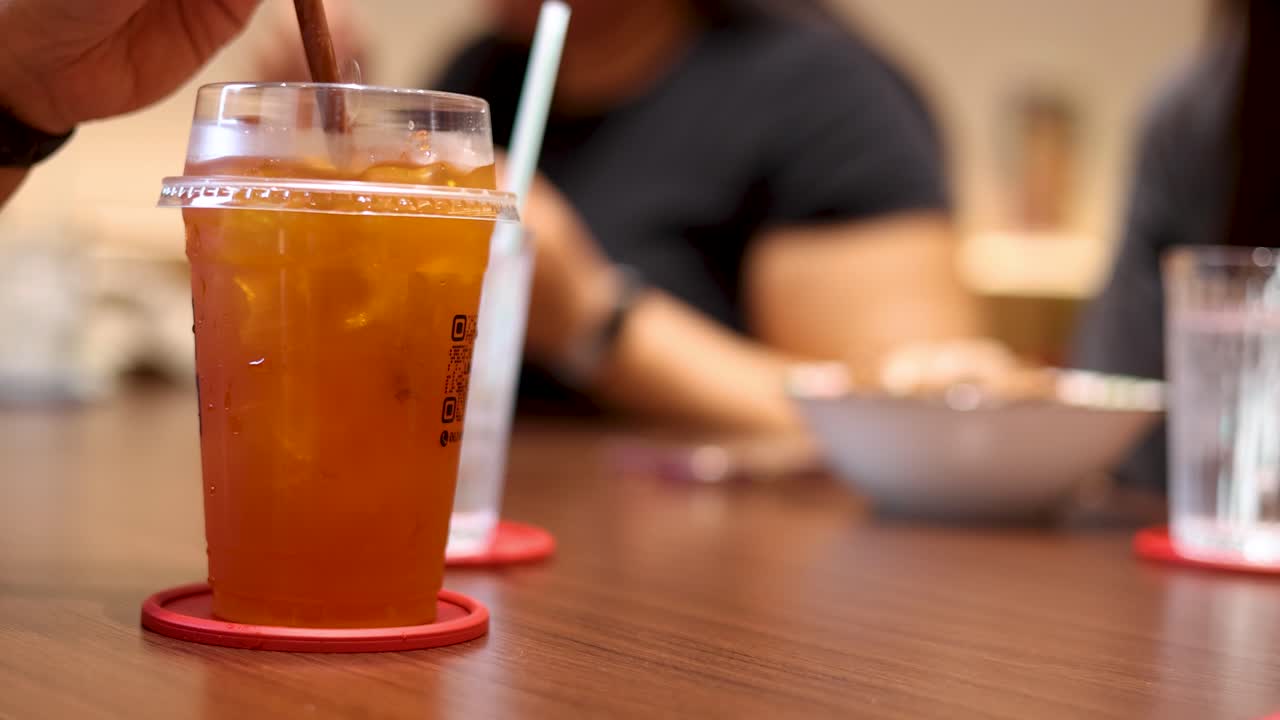 A hand stirs iced lemon tea in a clear plastic cup on a wooden table, with blurred background diners, warm indoor lighting, and shallow depth of field