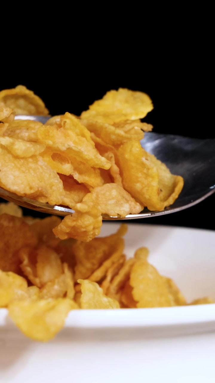 Un cucchiaio solleva i cornflakes da una ciotola bianca, su uno sfondo nero, evidenziando la consistenza del cereale.