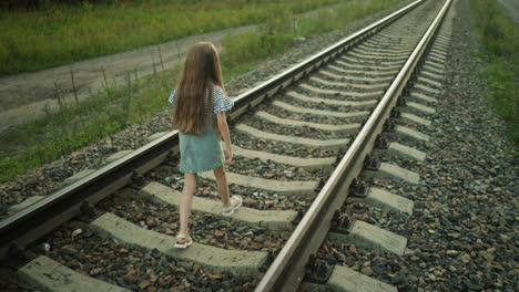 back view of kid with long hair walking on rail track in rural area, moment captured as she almost missteps onto gravel between sleepers, wearing denim dress and sandals, surrounded by greenery